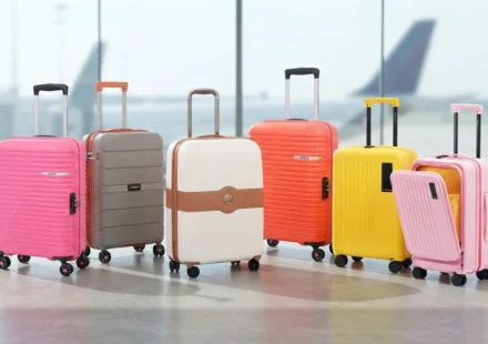 A collection of colorful hardside spinner luggage suitcases arranged neatly in a bright modern airport terminal with airplane views in the background.