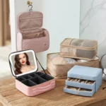 A stylish wooden dresser displaying various travel accessories including a pink hanging toiletry bag, LED makeup travel case, packing cubes, a blue jewelry box, and a neck pillow.