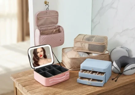 A stylish wooden dresser displaying various travel accessories including a pink hanging toiletry bag, LED makeup travel case, packing cubes, a blue jewelry box, and a neck pillow.